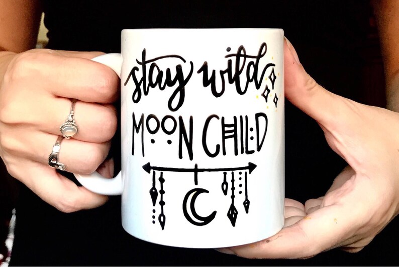 K&ouml;nnte beinhalten: Wei&szlig;e Keramiktasse mit schwarzer Aufschrift "stay wild moon child". Die Tasse hat einen schwarzen Halbmond und ein schwarzes Design aus h&auml;ngenden Pfeilen mit Punkten.