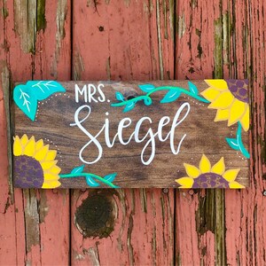 May include: A rectangular wooden sign with the name "Mrs. Siegel" in white script. The sign is decorated with yellow sunflowers with purple centers and teal leaves and vines. The background is a rustic brown.