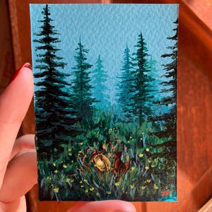 May include: Miniature painting of a forest scene with tall evergreen trees and a misty blue sky. The foreground features green grass and yellow flowers, with a glowing light source in the center. The artwork is on a small rectangular canvas.
