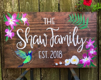 May include: Wooden family sign with the text "The Shaw Family EST. 2018" in white script. The sign is decorated with painted flowers, green leaves, and a hummingbird. The background is a stained wood.