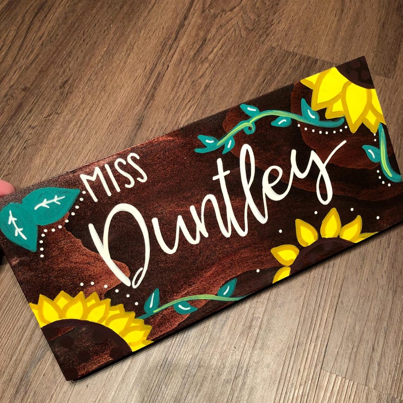 May include: A rectangular wooden sign with the name "Miss Duntley" in white script. The sign is decorated with yellow sunflowers, green vines, and teal leaves against a brown background.