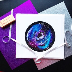 May include: A white card with a watercolor painting of a galaxy in shades of blue, purple, and white. The galaxy is swirling and has a dark center. The card is on a purple and silver glitter background.