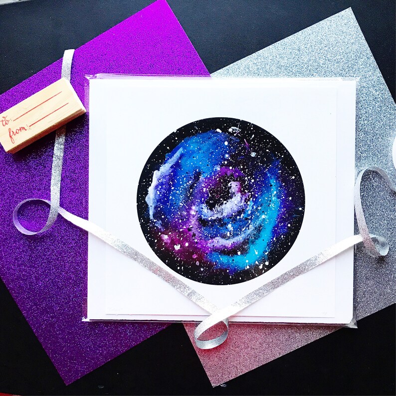 May include: A white card with a watercolor painting of a galaxy in shades of blue, purple, and white. The galaxy is swirling and has a dark center. The card is on a purple and silver glitter background.