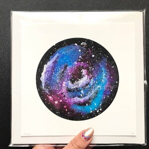 May include: A watercolor painting of a galaxy in shades of blue, purple, and white on a black background. The galaxy is surrounded by a white border and the painting is framed in a white mat.