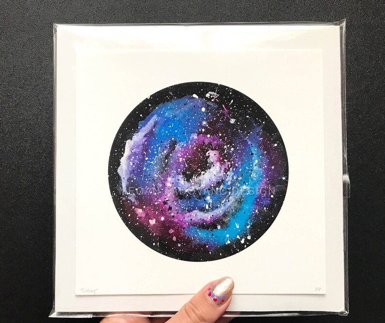 May include: A watercolor painting of a galaxy in shades of blue, purple, and white on a black background. The galaxy is surrounded by a white border and the painting is framed in a white mat.