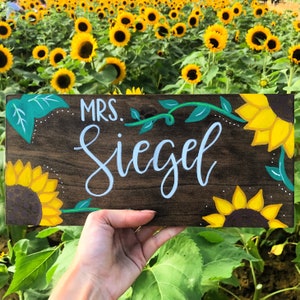 May include: A wooden sign with the name "Mrs. Siegel" in light blue script, decorated with yellow sunflowers and green vines. The sign is set against a backdrop of a field of sunflowers.