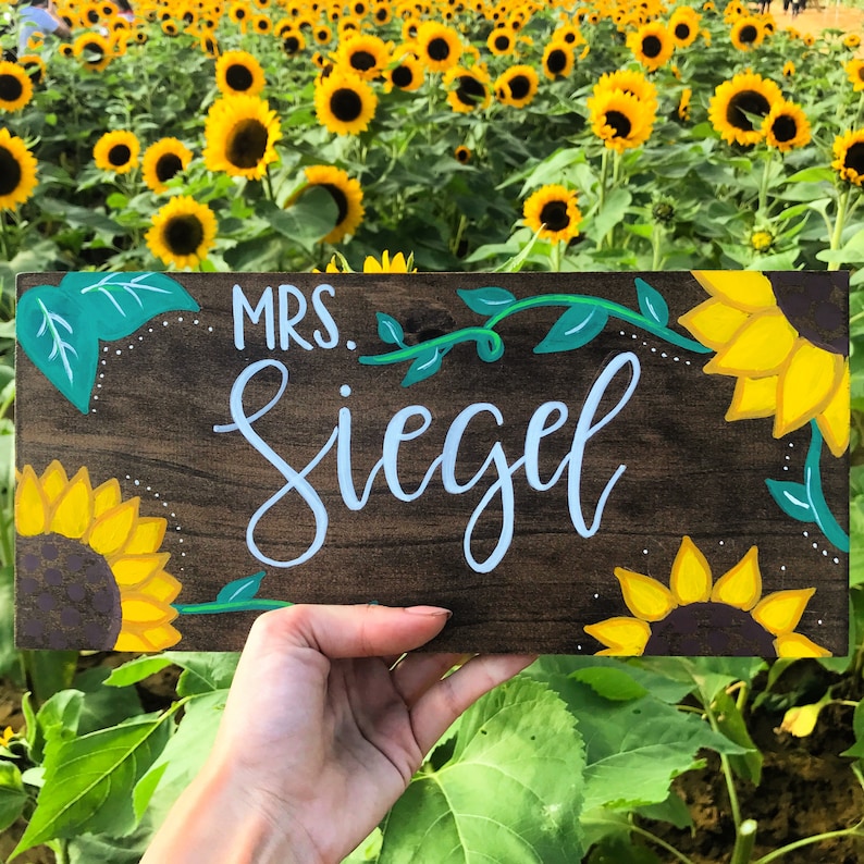 May include: A wooden sign with the name "Mrs. Siegel" in light blue script, decorated with yellow sunflowers and green vines. The sign is set against a backdrop of a field of sunflowers.