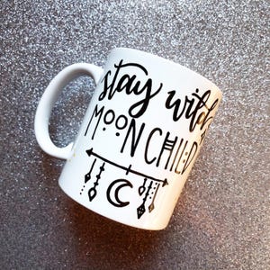 A white mug with black text and decorative elements, such as a crescent moon and stars, on a glittery surface.