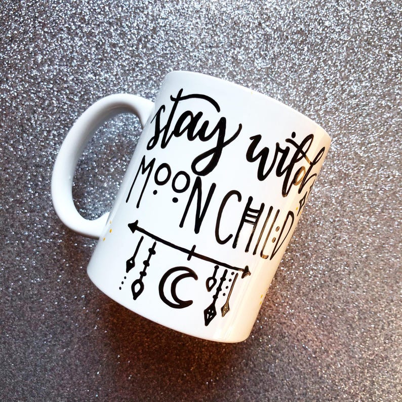 A white mug with black text and decorative elements, such as a crescent moon and stars, on a glittery surface.