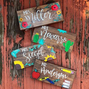 May include: Four wooden teacher name signs with painted floral and school-themed designs. The signs are personalized with the names "Mrs. Miller", "Mrs. Navarro", "Mrs. Siegel", and "Mr. Anderson". The signs are on a rustic wooden background.