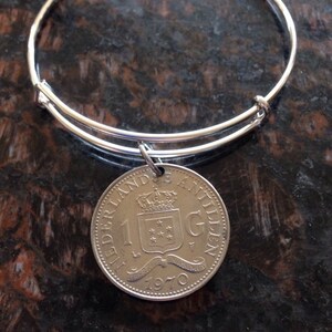 May include: A silver bangle bracelet with a silver charm featuring a 1970 Dutch Guilder coin. The coin has a coat of arms with a crown and stars.