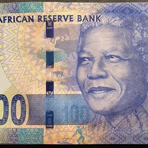 May include: A South African 100 Rand banknote featuring a portrait of Nelson Mandela on the right side. The bill displays the text South African Reserve Bank, One Hundred Rand, and the number 100 in large blue and green metallic ink fonts.