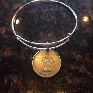 Könnte beinhalten: Ein silbernes Armreif-Armband mit einem goldfarbenen Münzenanhänger, der ein traditionelles arabisches Kaffeekännchen zeigt und die Inschrift "1990-1910" trägt.