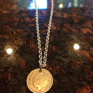 Könnte beinhalten: Eine silberne Kettenkette mit einem goldenen Münzenanhänger, der das Profil einer Person zeigt. Die Münze hat einen erhöhten Rand und ist leicht abgenutzt.