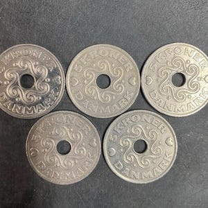 May include: Five silver-colored Danish 5 Kroner coins arranged in a group. Each circular coin features a central hole, a swirling decorative pattern, and the text 5 KRONER DANMARK along the outer edge, accompanied by small heart symbols.