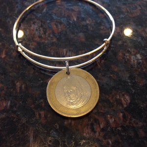 Könnte beinhalten: Ein goldenes Armreif-Armband mit einem runden Münzenanhänger, der ein Porträt zeigt. Die Münze ist silber- und goldfarben.