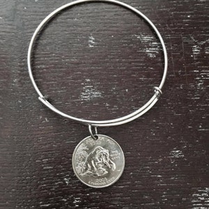 May include: A silver bangle bracelet with a 2009 Alaska quarter charm featuring a brown bear and the text "The Great Land".
