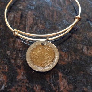 Könnte beinhalten: Ein goldenes Armreif-Armband mit einem silbernen Münzenanhänger, der ein Vogelmotiv zeigt. Die Münze trägt die Inschriften "Republik der Philippinen" und "1 Peso".