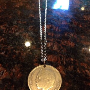 Könnte beinhalten: Eine silberne Kettenkette mit einem goldenen Münzenanhänger, der ein Profilporträt eines Mannes zeigt. Die Münze hat arabische Schrift am Rand.