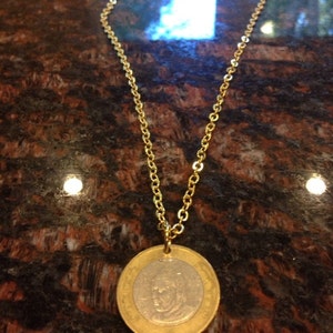 Könnte beinhalten: Eine goldene Kettenkette mit einem runden Münzenanhänger, der das Profil einer Person zeigt. Die Münze ist zweifarbig, mit einem goldenen Außenring und einer silbernen Mitte.