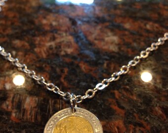 Könnte beinhalten: Eine silberne Kettenkette mit einem goldenen Münzenanhänger mit einem detaillierten Design. Die Münze ist eine mexikanische 5-Peso-Münze.
