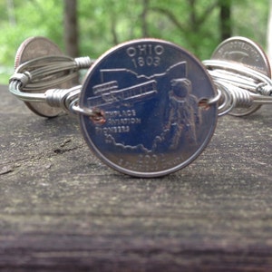 Peut inclure: Un bracelet en argent fait de fil et de pièces de monnaie américaines de 25 cents. La pièce de 25 cents sur le bracelet porte le texte "Ohio 1803" et une image d'un avion. Le texte "Birthplace Aviation Pioneers" est également visible sur la pièce.
