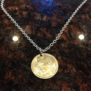 Könnte beinhalten: Eine silberne Kettenkette mit einem runden Münzenanhänger, der ein Profilporträt eines Mannes zeigt. Die Münze ist goldfarben und hat einen erhöhten Rand.