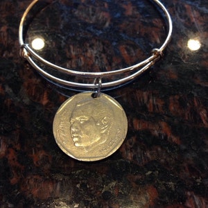 Könnte beinhalten: Ein silbernes Armreif-Armband mit einem Münzenanhänger, der das Profil eines Mannes zeigt. Die Münze ist goldfarben und hat ein erhabenes Design.