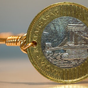 May include: A gold-toned wire bracelet featuring a Hungarian coin as a focal point. The coin has a detailed image of a bridge and is secured with wrapped wire. The bracelet's design is simple yet elegant.