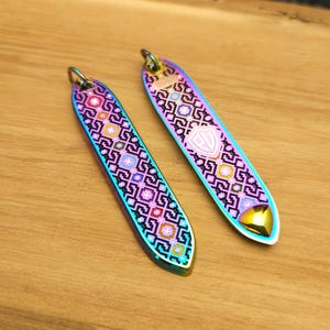 Puede incluir: Dos colgantes metálicos alargados e iridiscentes con intrincados patrones geométricos en rosa, morado y turquesa. Cada colgante tiene un pequeño lazo en la parte superior y una punta redondeada de color dorado. Los colgantes se muestran sobre una superficie de madera.
