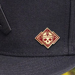 May include: Black baseball cap with a black embroidered letter 'A' on the front and a gold and red enamel pin with a skull wearing a crown on the side.