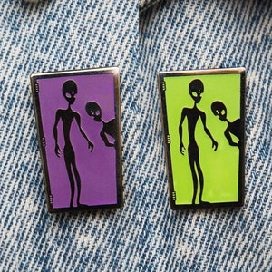 May include: Two silver pins with black silhouettes of aliens inside a rectangle. The pin on the left has a purple background and the pin on the right has a green background.