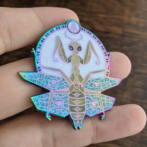 May include: A iridescent green praying mantis with wings outstretched, surrounded by a circle of glitter and a small tree symbol. The mantis is set against a teal and purple background with a geometric pattern.