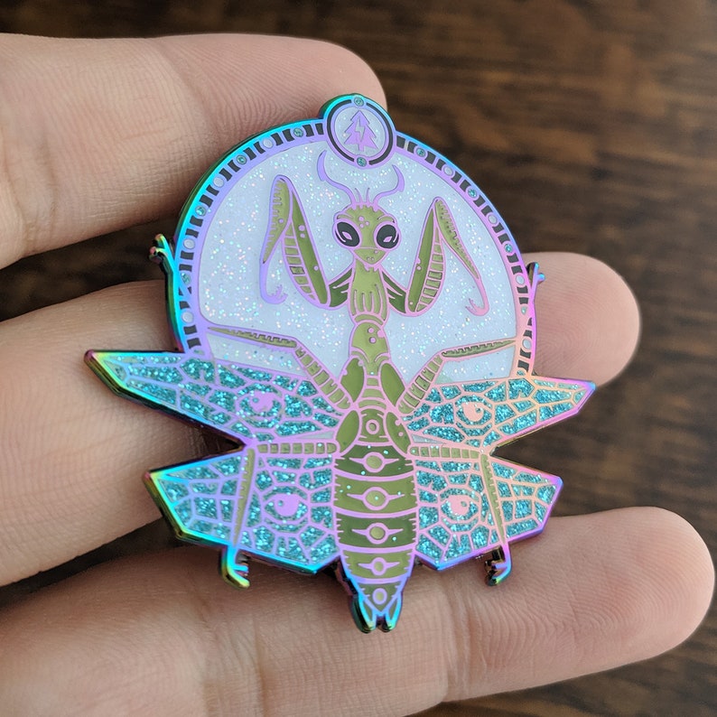 May include: A iridescent green praying mantis with wings outstretched, surrounded by a circle of glitter and a small tree symbol. The mantis is set against a teal and purple background with a geometric pattern.