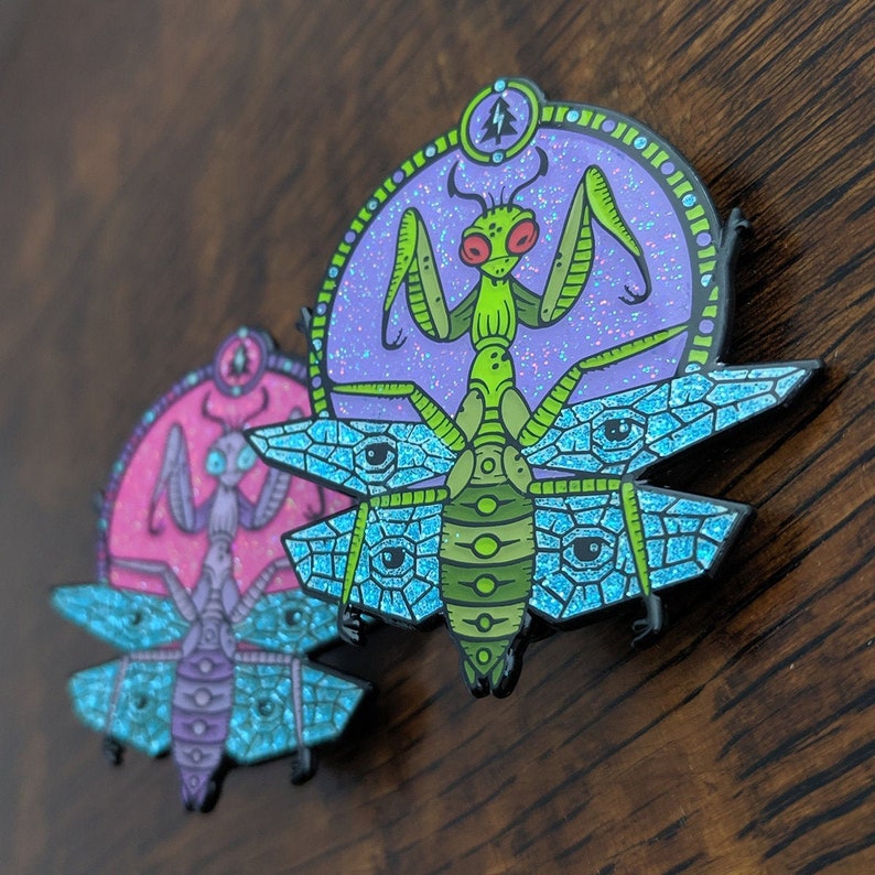 May include: Two enamel pins featuring praying mantises with intricate details. The top pin has a green mantis with blue and purple wings and a purple background. The bottom pin has a pink mantis with blue and purple wings and a pink background.
