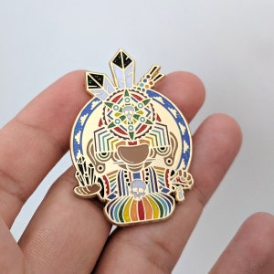 Puede incluir: Pin de esmalte dorado con un diseño geométrico colorido con una calavera y plumas. El pin representa una figura estilizada con una túnica de colores arcoíris y una calavera en el pecho.