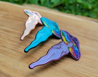 Puede incluir: Tres pines de esmalte con forma de figura desnuda con un sombrero de hongo. Los pines son en tonos cobre, turquesa y morado con detalles arcoíris e iridiscentes. Los pines están sobre una superficie de madera.