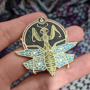 May include: Gold-tone enamel pin featuring a green praying mantis with a blue and green geometric pattern behind it. The mantis is surrounded by a circle with a black background and gold glitter.