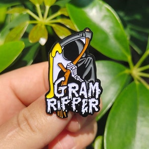 Puede incluir: Pin de esmalte negro y amarillo con un esqueleto sosteniendo una guadaña con el texto "Gram Ripper" en blanco.