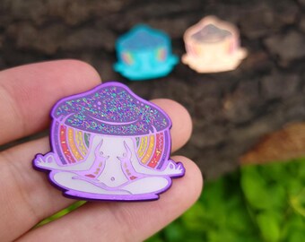 Puede incluir: Un pin de esmalte morado que representa una figura meditando con una cabeza de hongo. La tapa del hongo está cubierta de purpurina. El cuerpo de la figura es blanco con detalles de arcoíris. Hay otros dos pines en el fondo.