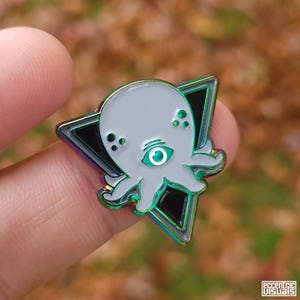 May include: A gray octopus enamel pin with a single green eye, set within a black triangle with iridescent edges. The pin is held between fingers against a blurred background.