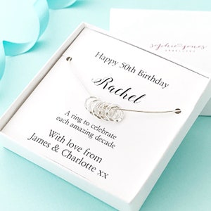 May include: A white gift box with a silver necklace inside. The necklace has ten small silver rings, each representing a decade. The box has a personalized message that reads "Happy 50th Birthday Rachel. A ring to celebrate each amazing decade. With love from James & Charlotte xx."