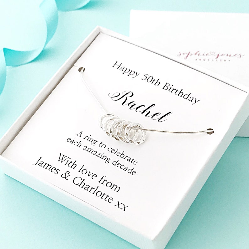 May include: A white gift box with a silver necklace inside. The necklace has ten small silver rings, each representing a decade. The box has a personalized message that reads "Happy 50th Birthday Rachel. A ring to celebrate each amazing decade. With love from James & Charlotte xx."