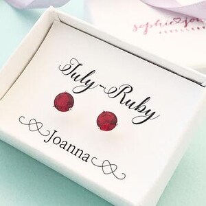 May include: A white gift box containing a pair of ruby red stud earrings. The box is printed with the words "July-Ruby" and "Joanna" in a decorative script. The earrings are circular and likely set in silver or white metal.