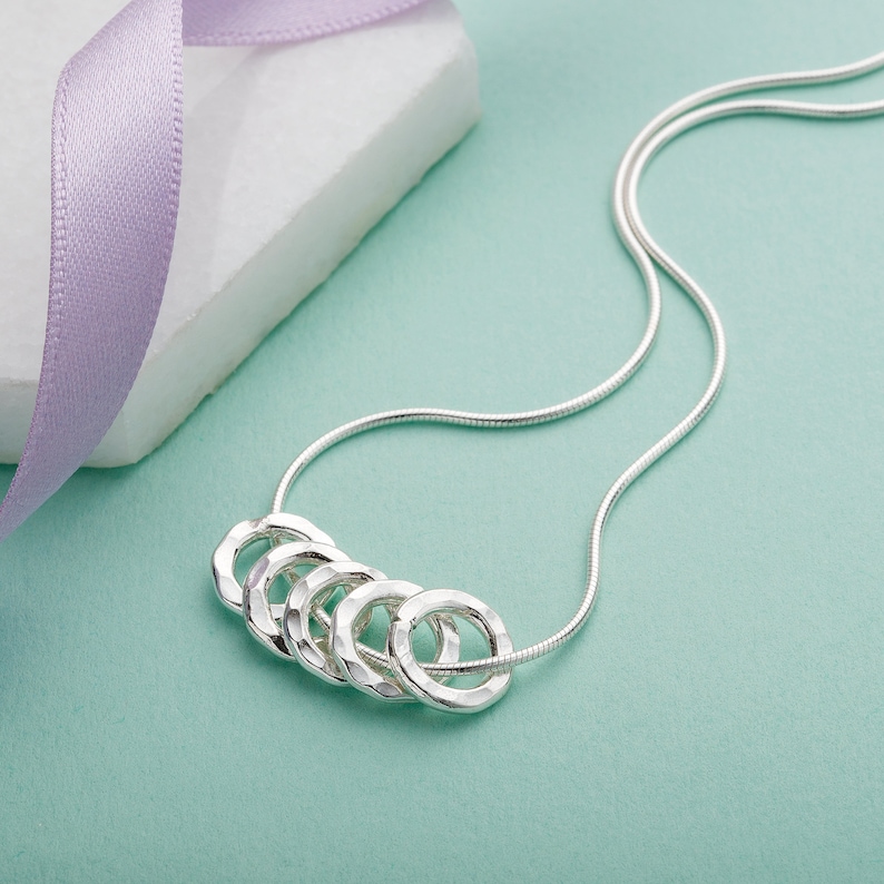 May include: A silver necklace with five interconnected hammered rings. The necklace is on a light blue background.