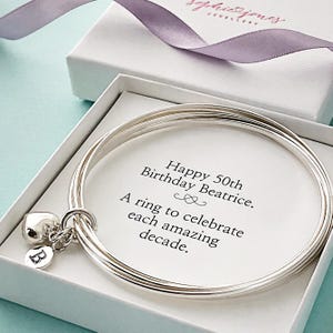 May include: A silver bangle bracelet in a white gift box, with a heart charm and a disc charm with the letter "B". The card inside reads "Happy 50th Birthday Beatrice. A ring to celebrate each amazing decade."