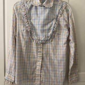 May include: A long-sleeved plaid shirt in beige, light blue, and white. The shirt features a ruffled collar and bib, with small silver-colored buttons down the front and on the cuffs. The fabric has a vintage aesthetic.