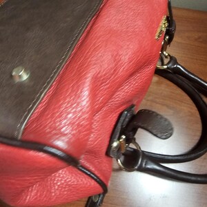 May include: A handbag featuring a textured red leather body with dark brown leather accents and trim. The bag includes dark brown leather handles, gold-tone metal hardware, and a small gold-tone logo emblem attached to the upper side panel.