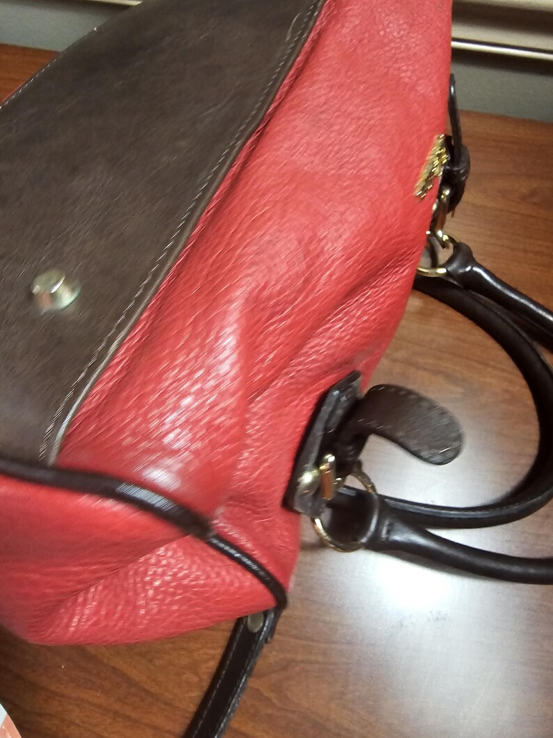 May include: A handbag featuring a textured red leather body with dark brown leather accents and trim. The bag includes dark brown leather handles, gold-tone metal hardware, and a small gold-tone logo emblem attached to the upper side panel.