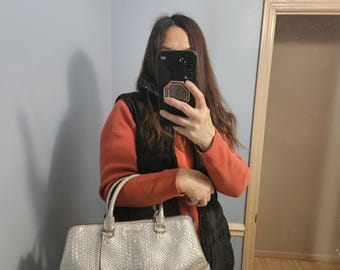 May include: A silver-colored handbag with a snakeskin pattern. The bag has two handles and a gold-colored circular charm with the brand's initials. The bag is held by a person in front of a mirror.